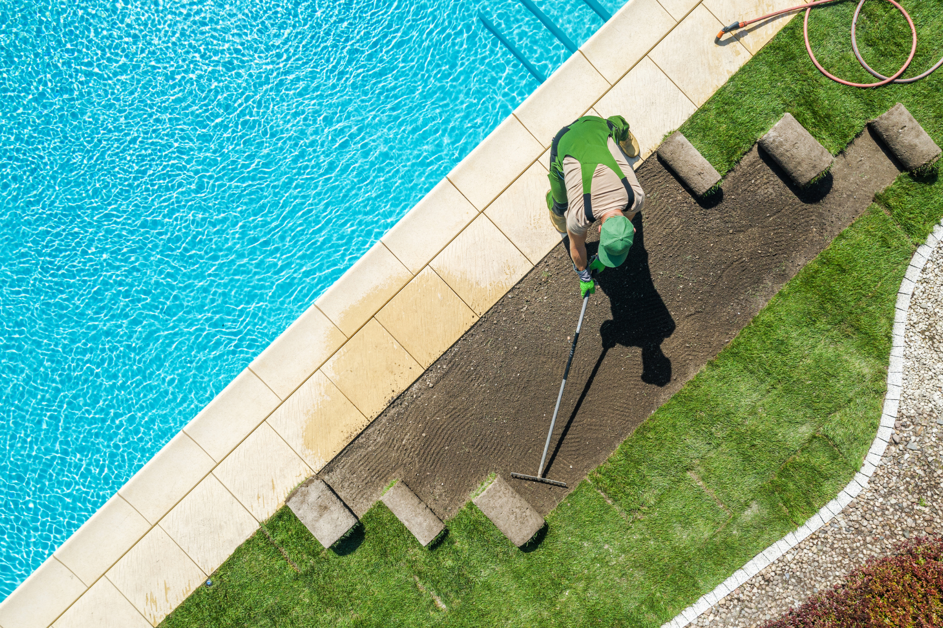 Person using a string trimmer beside a pool on a green lawn and stone path