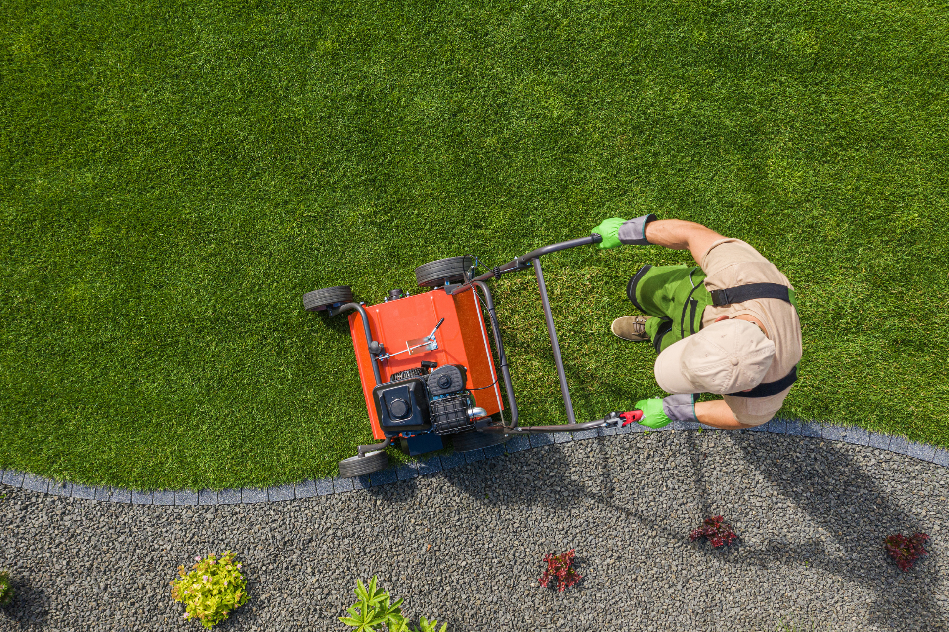 Person operating a red lawn mower on green grass beside a gravel edge.