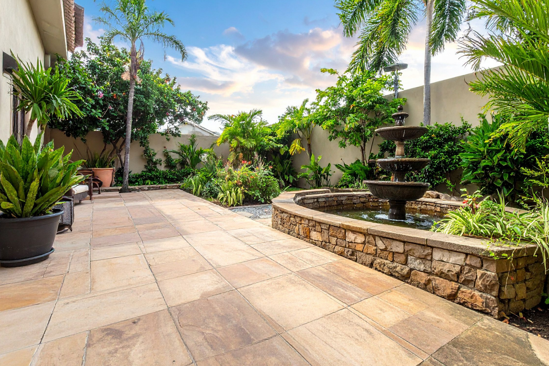 Courtyard patio with stone fountain, potted plants, and lush greenery under a bright sky