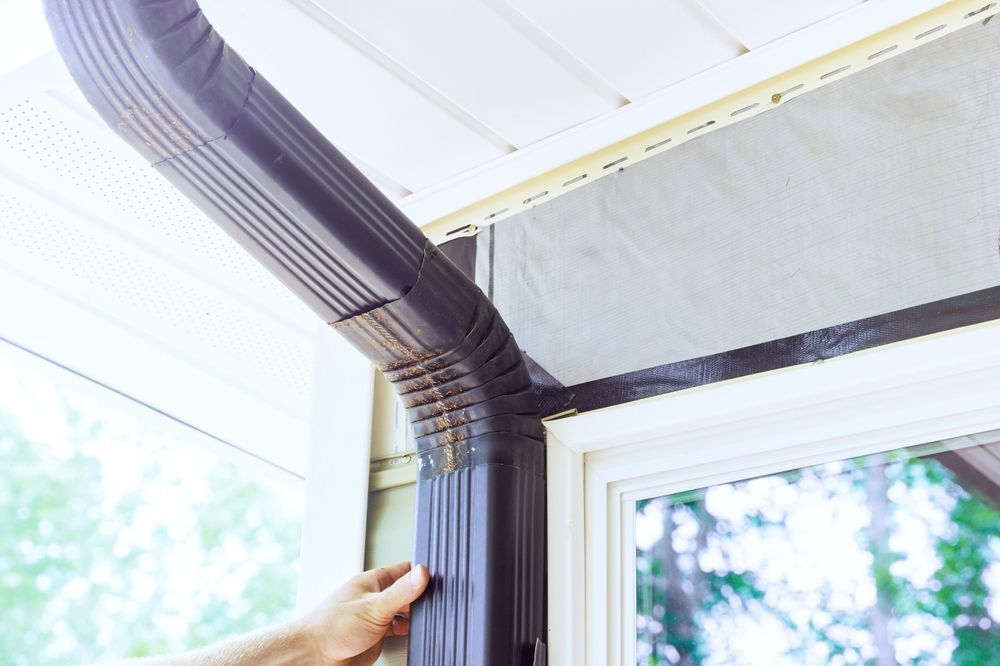 Hand installing a brown gutter downspout at a roof corner beside a white house window