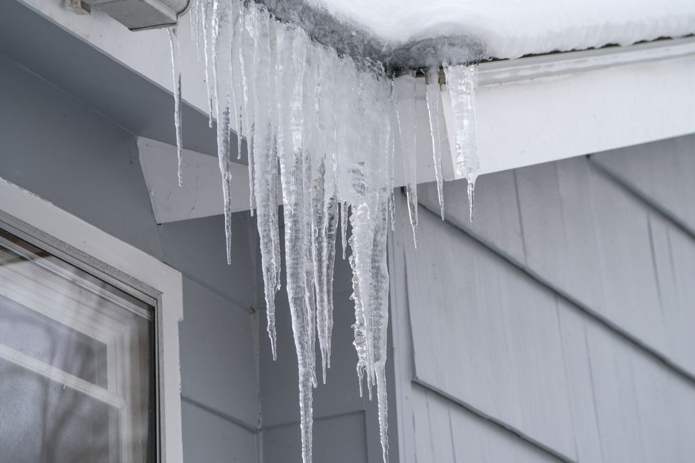 Icicles hanging from a house roof over gray siding and a window