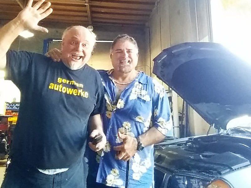 Two smiling people standing by an open car hood in a garage, one waving