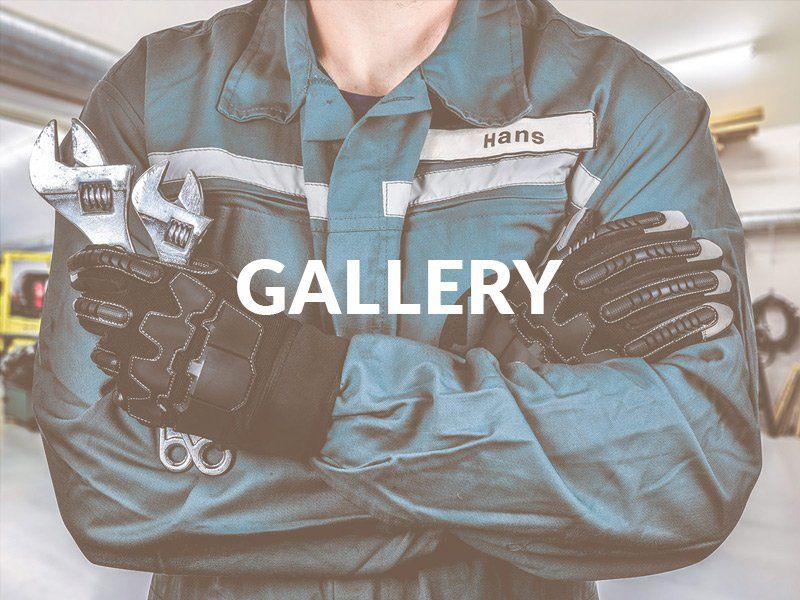 Person in teal work jacket wearing black gloves with “GALLERY” text overlay in a workshop setting