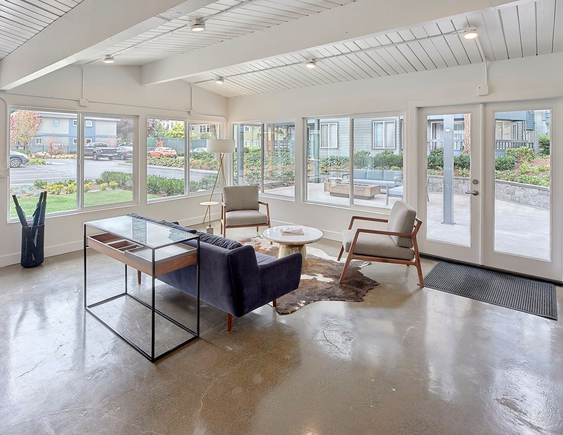 A sunlit, minimalist lobby with a blue sofa, armchairs, a glass console table, and large windows on a polished floor.