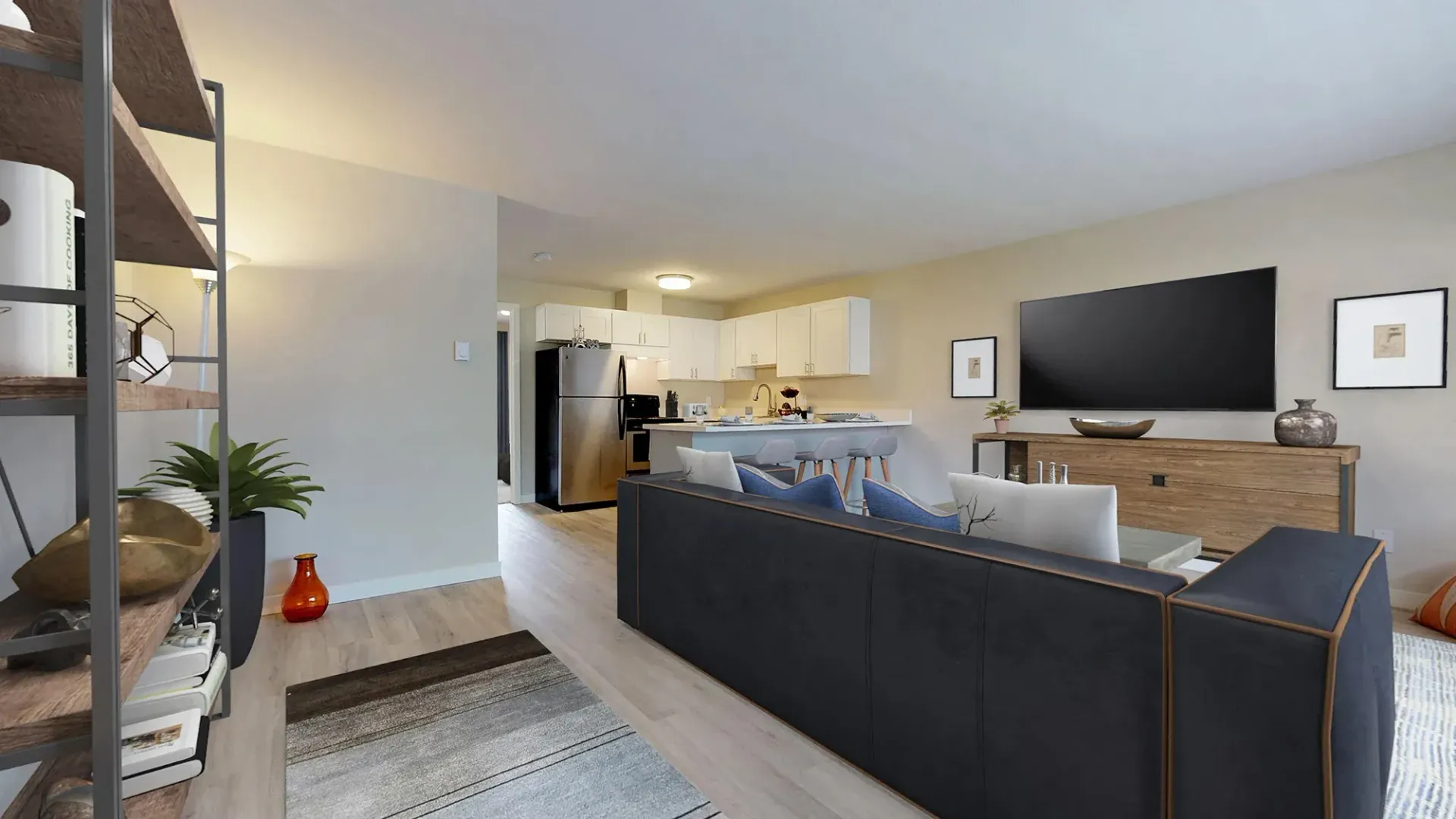 A modern, open-concept apartment living room featuring a dark gray sofa, light wood floors, and a kitchen in the back.