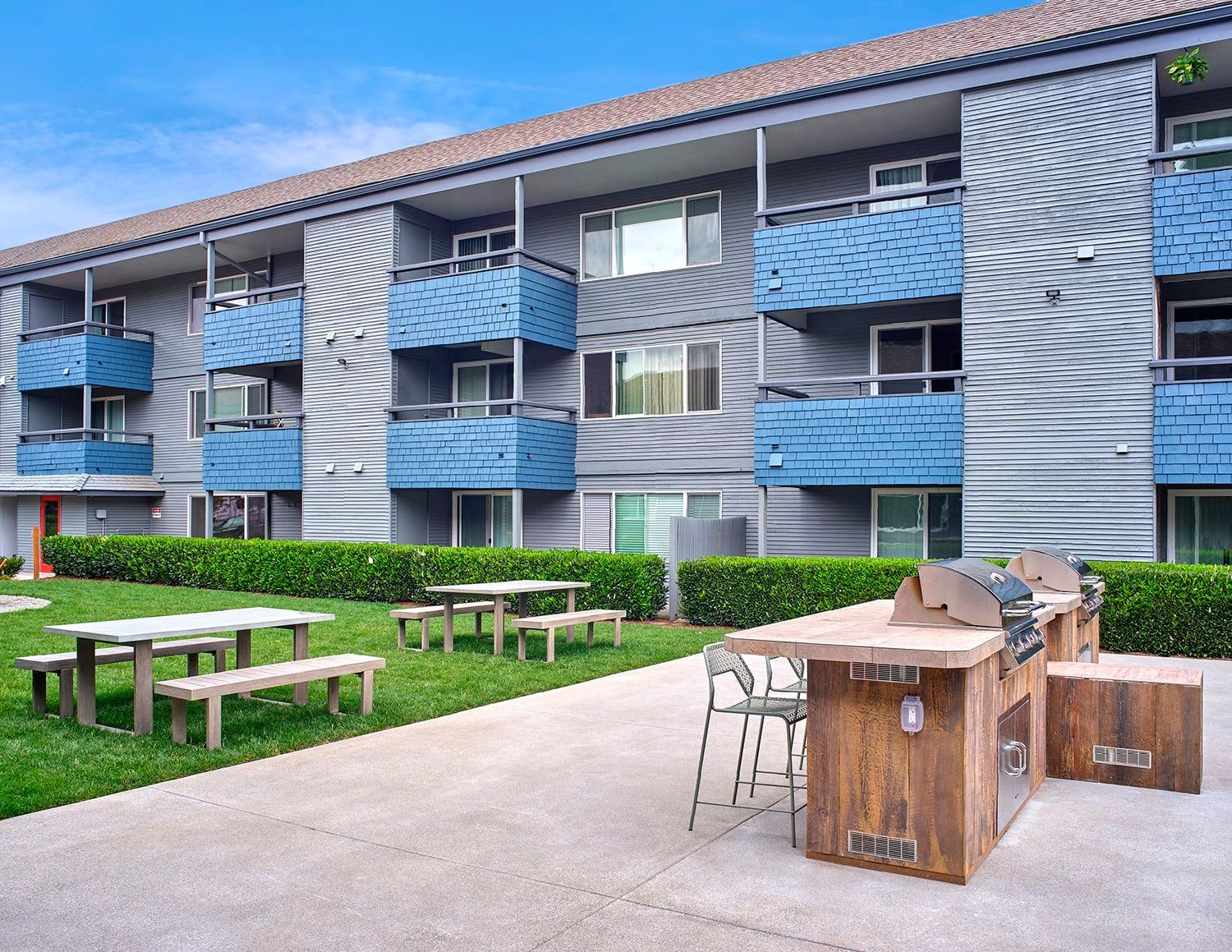 A three-story apartment complex with blue balconies, featuring an outdoor grill area and picnic tables on a lawn.