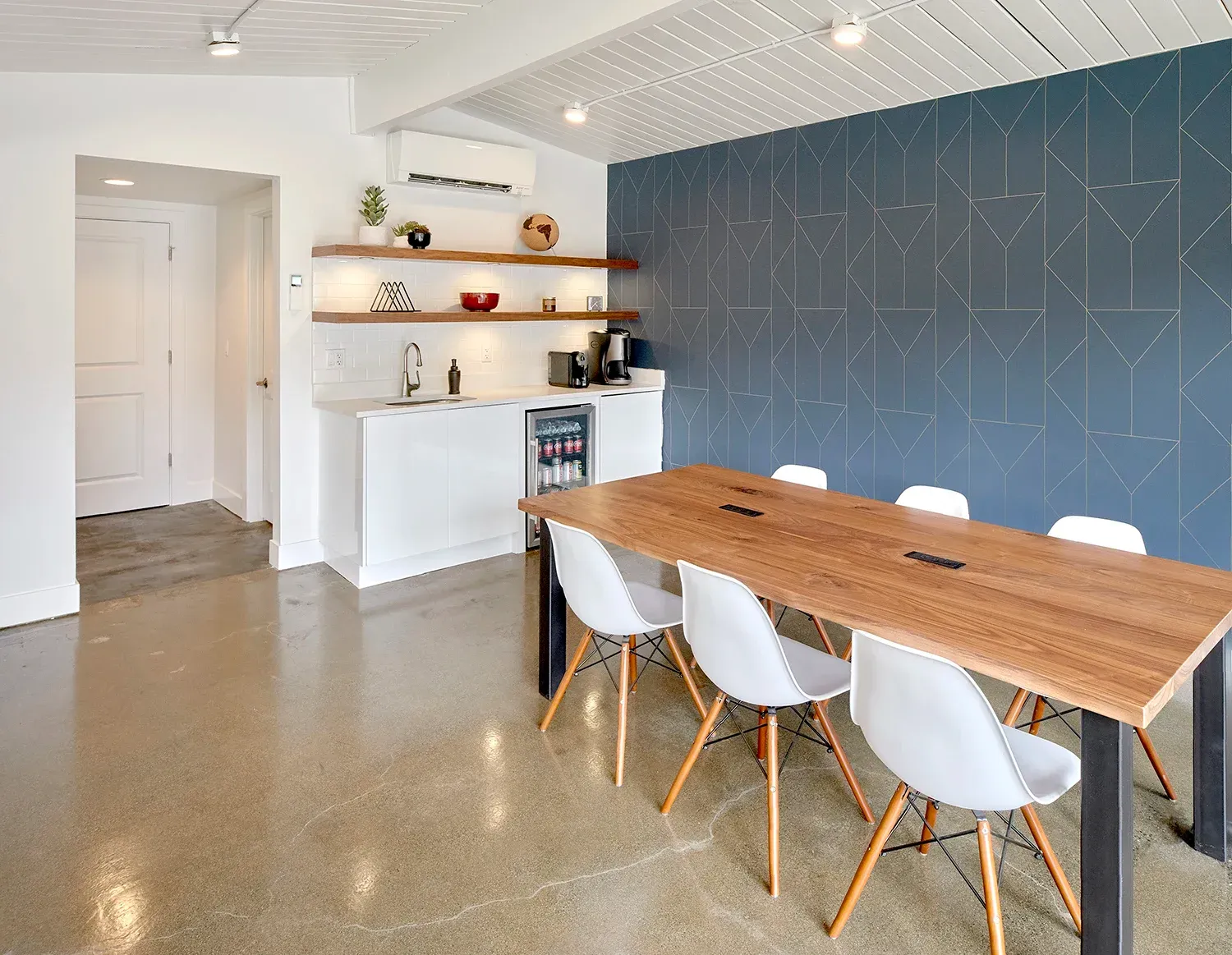 A modern meeting room features a long wooden table with six white chairs, set against a geometric navy blue accent wall.
