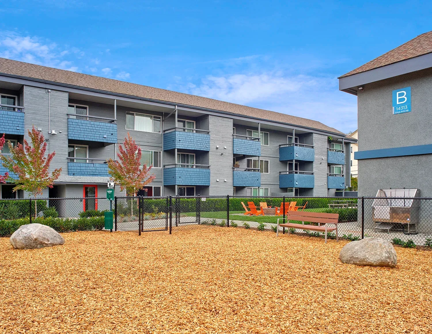A dog park with wood chips, two large rocks, a wooden bench, and an apartment building in the background.
