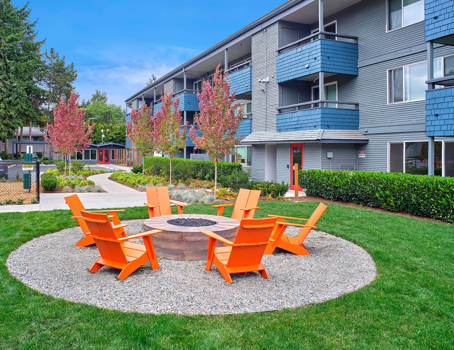 Bright orange Adirondack chairs arranged in a circle around a stone fire pit on a gravel patio beside an apartment building.