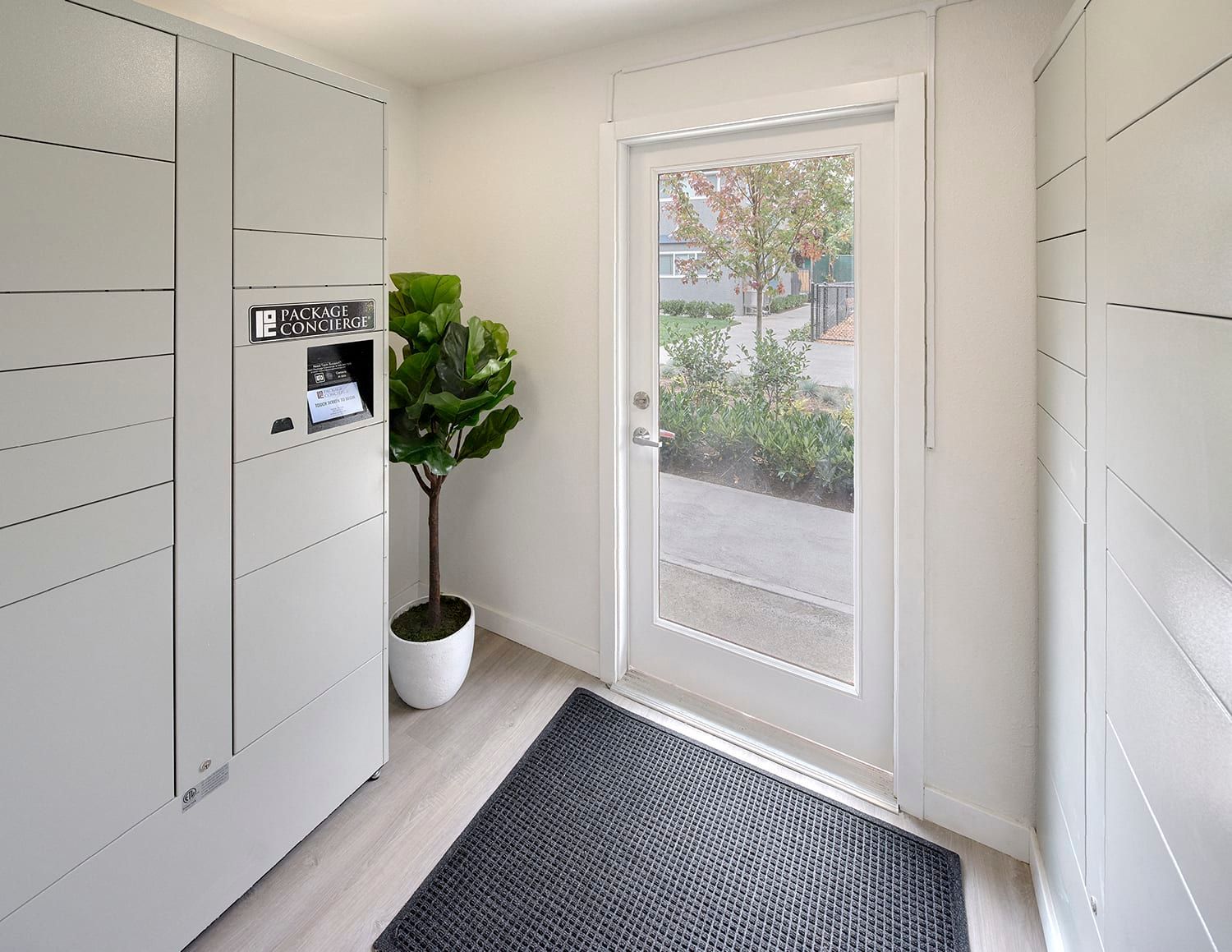 A white parcel locker system stands in a bright entrance area next to a glass-paneled door, a potted plant, and a mat.