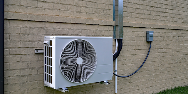Outdoor wall-mounted air conditioner unit attached to a beige brick building with visible wiring.