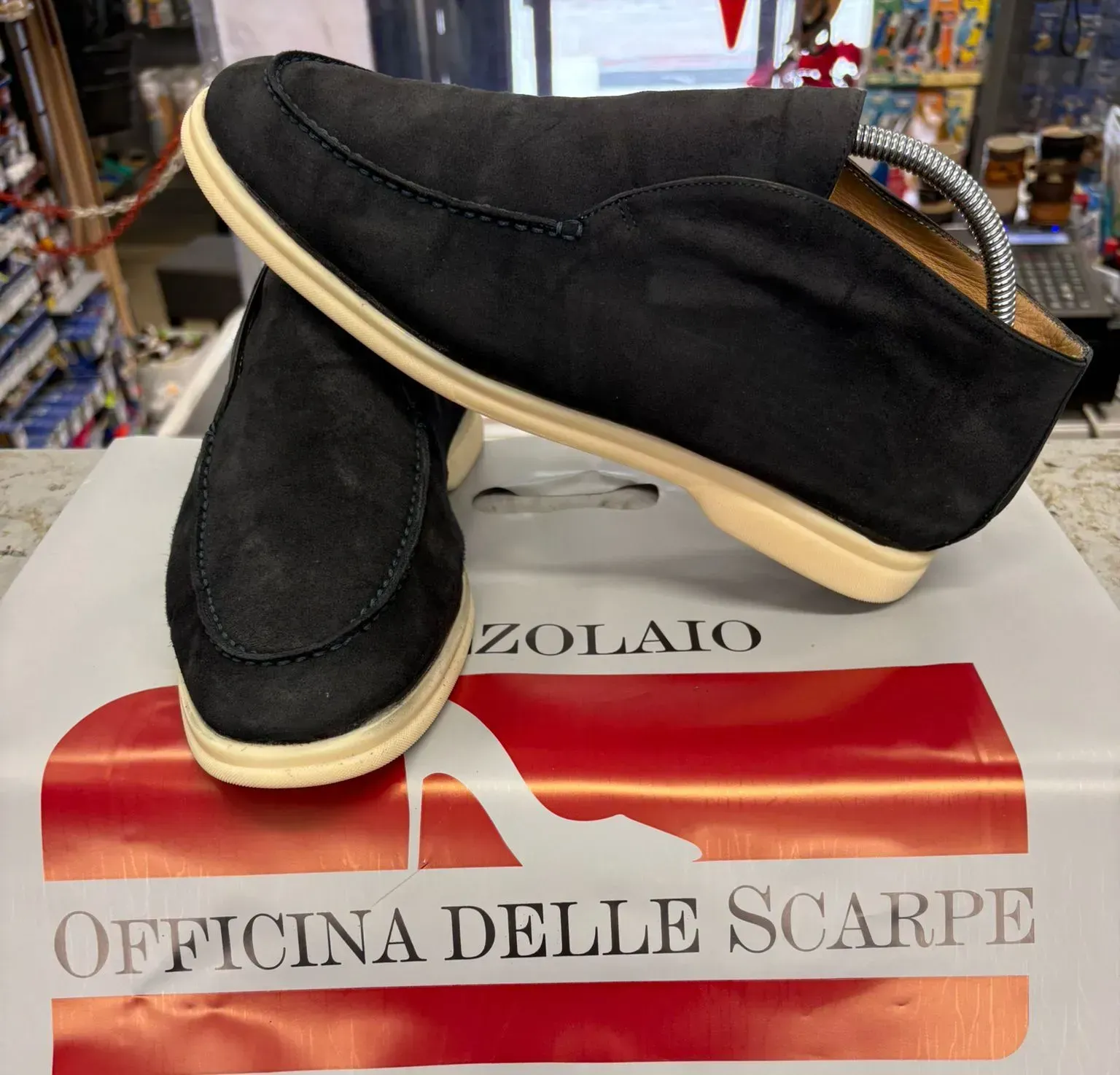 Scarpe slip-on nere con suola beige esposte su un cartello ZOLAIO Officina delle Scarpe.
