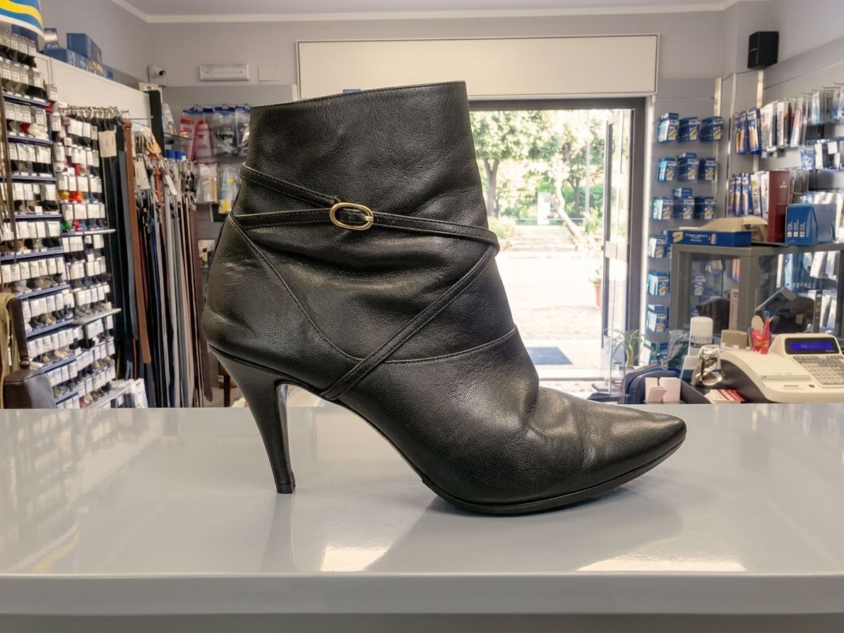 Stivaletto alla caviglia in camoscio nero con tacco alto, punta affusolata e fibbia dorata, esposto in un negozio di scarpe.