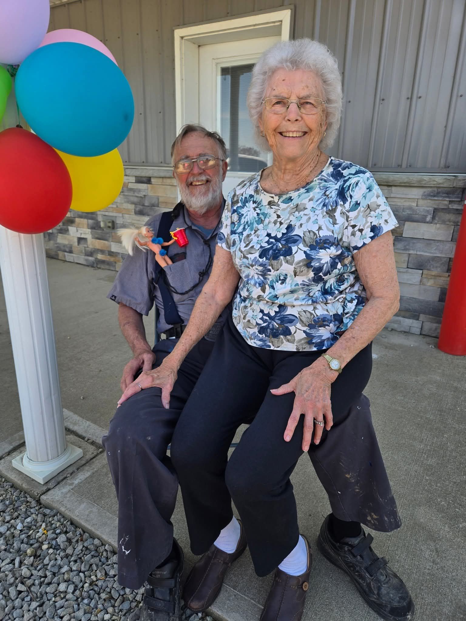 A person in a patterned top sits on the lap of another person in a gray shirt, both smiling next to colorful balloons.