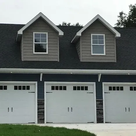Three white garage doors beneath gray dormer windows on a house with a dark shingled roof.