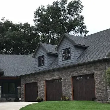 Stone house with three dark wood garage doors and dormer windows under gray roofs, framed by trees