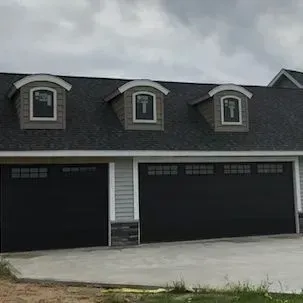 Three-car garage with dark doors and dormer windows on a gray house under an overcast sky.