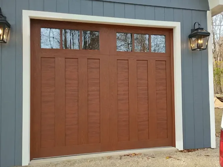 Wooden garage door with window panels set in a gray exterior wall, flanked by black lantern lights.