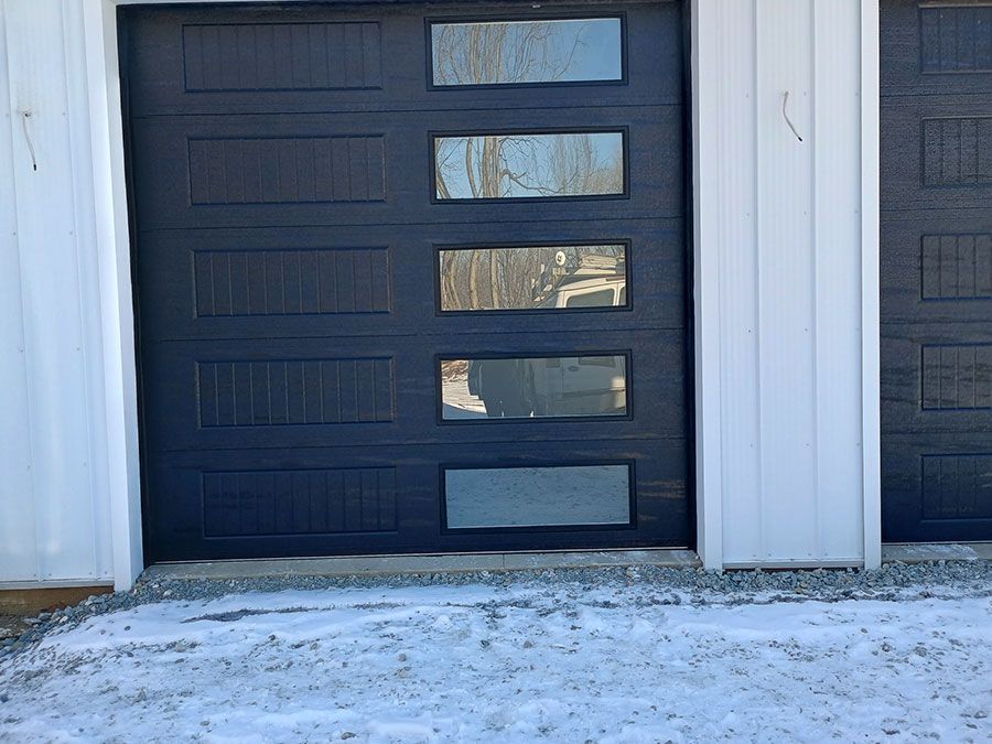 Black garage door with narrow windows beside white siding and snow-covered ground