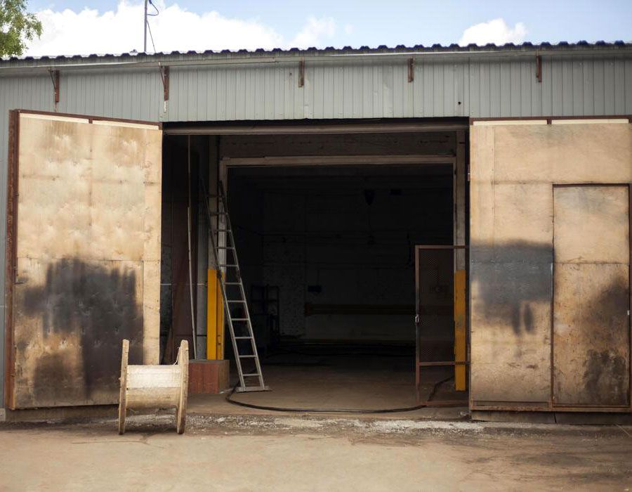 Open industrial warehouse with large metal doors, ladder inside, and a wheelbarrow outside.