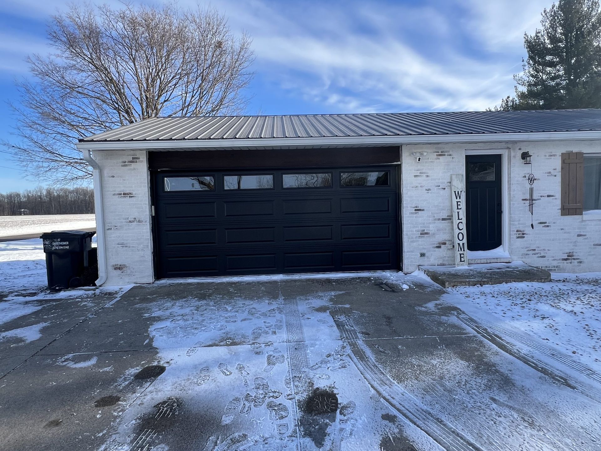 Snow-covered driveway in front of a house with a black garage door and white brick exterior.