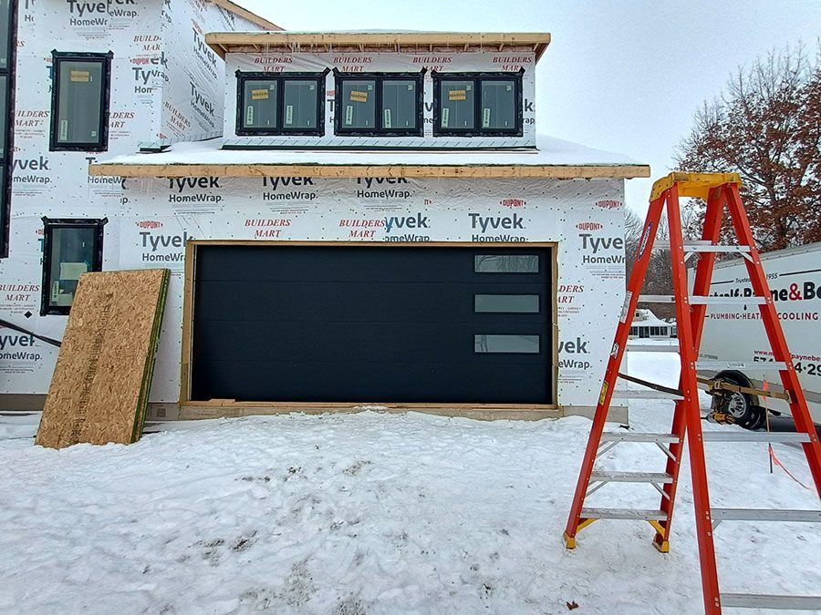 House under construction with black garage door, ladders, and snow on the ground