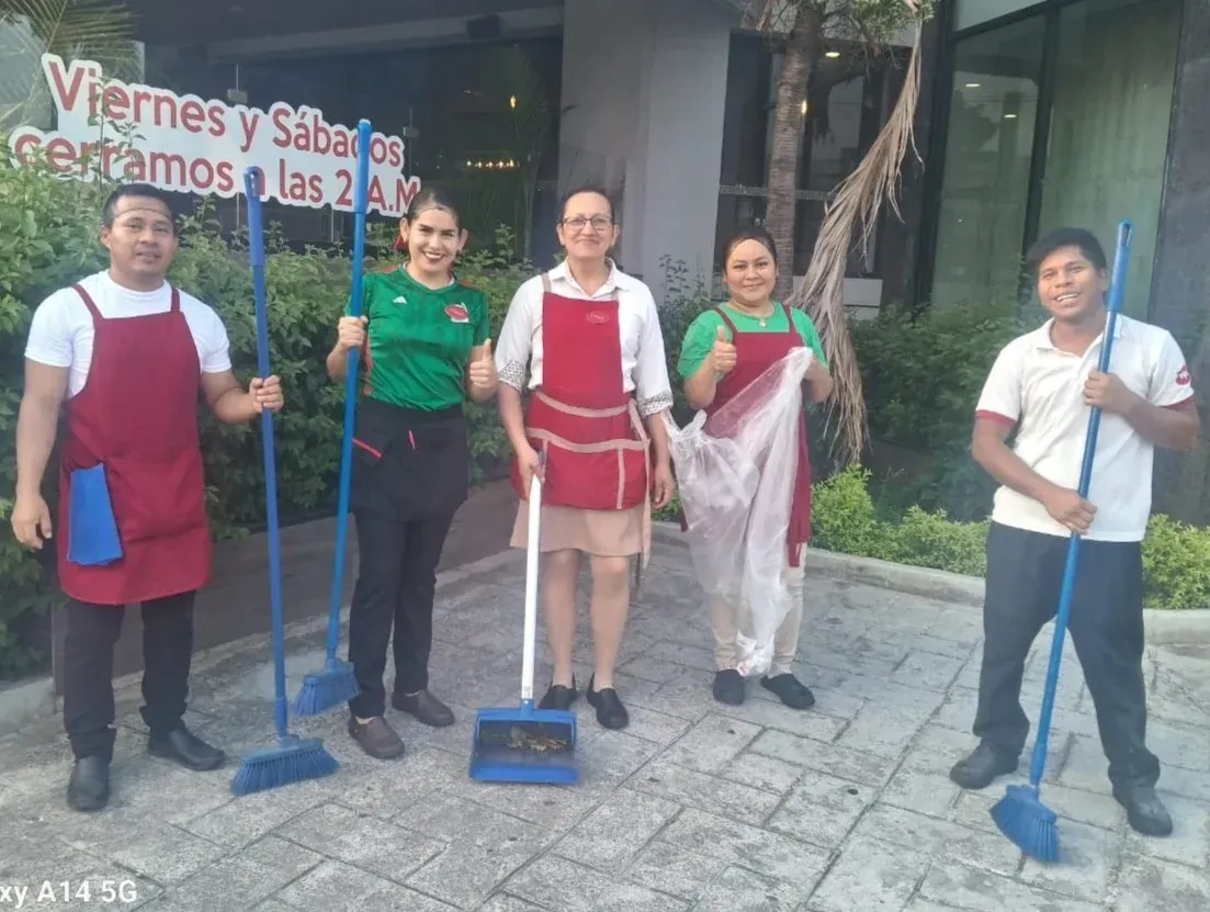 Cinco personas con delantales sostienen escobas y fregonas frente a un edificio, posando en una escena de limpieza.