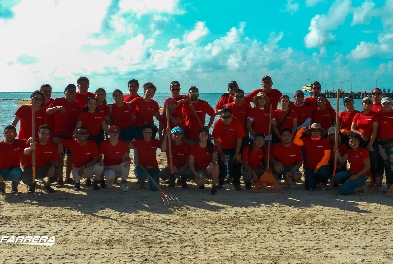 Un grupo numeroso de personas con camisetas rojas posando en una playa de arena bajo un cielo azul brillante.