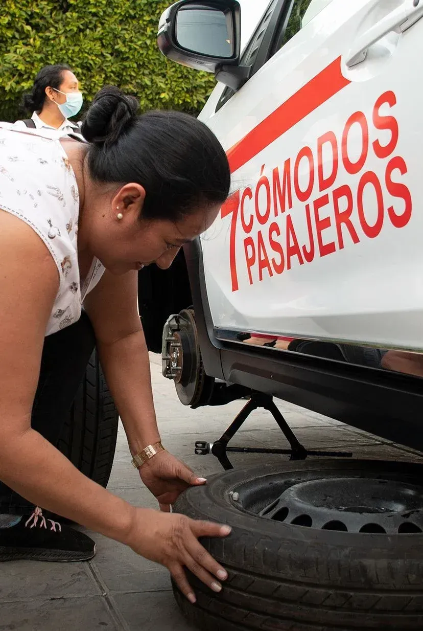 Mujer cambiando una llanta junto a un vehículo blanco de transporte de pasajeros con la etiqueta “TÓMONOS PASAJEROS”.