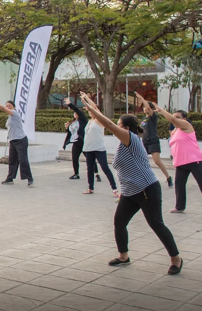 Gente haciendo ejercicio al aire libre en una plaza, estirándose con los brazos en alto cerca de una pancarta.