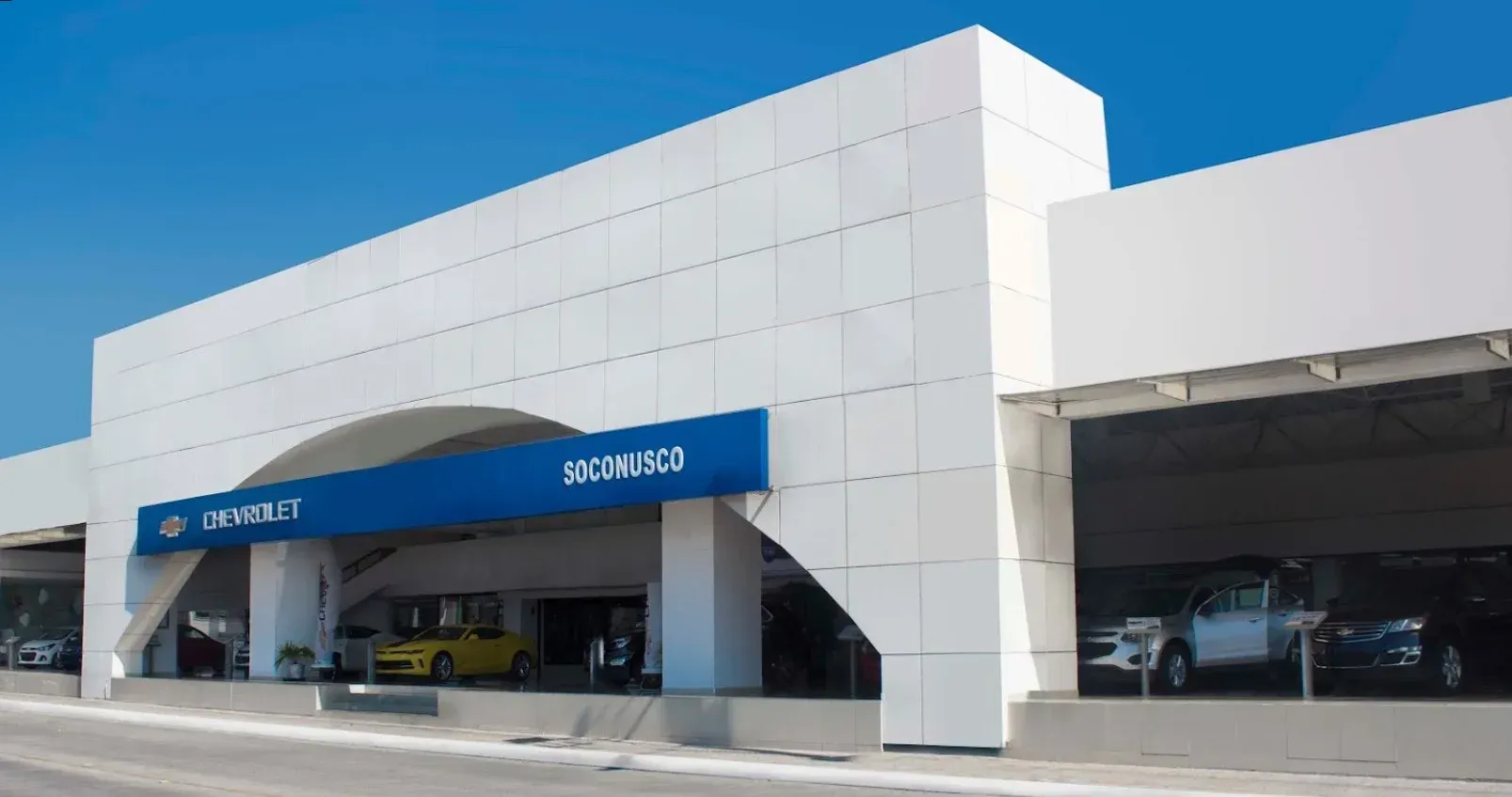 Exterior de un moderno edificio blanco con toldos azules y coches aparcados en un concesionario de automóviles.