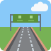 Camino que lleva a una señal de autopista verde bajo un cielo azul con nubes.