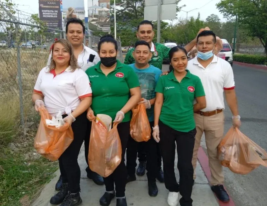 Voluntarios con camisetas verdes transportando bolsas de basura a lo largo de una carretera durante una jornada de limpieza.