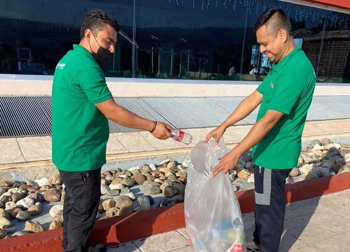Dos personas con camisetas verdes pasan objetos a una bolsa transparente junto a un edificio y unas rocas.