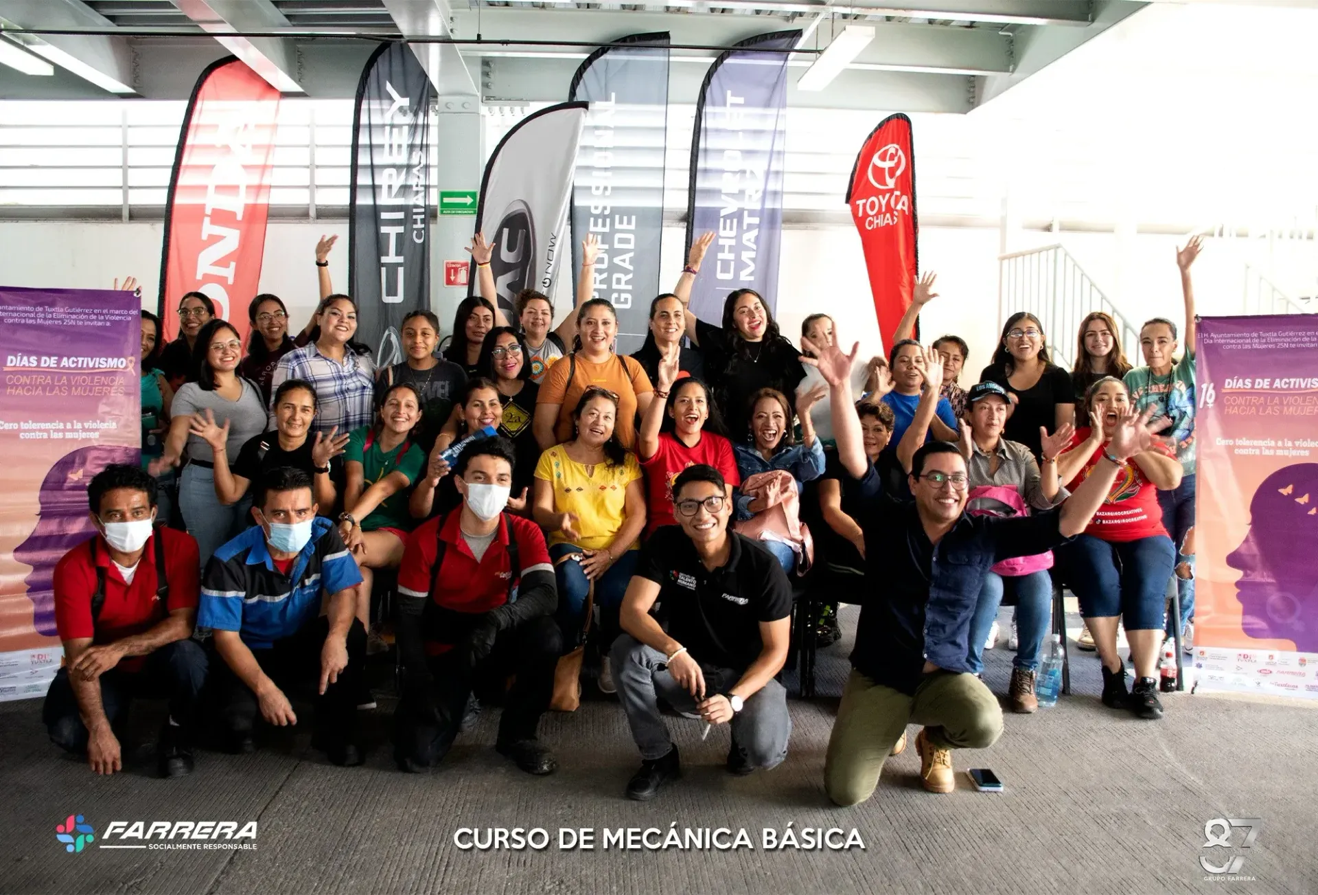 Foto grupal de personas sonrientes posando en interiores durante un evento de clase con pancartas y banderas de fondo.
