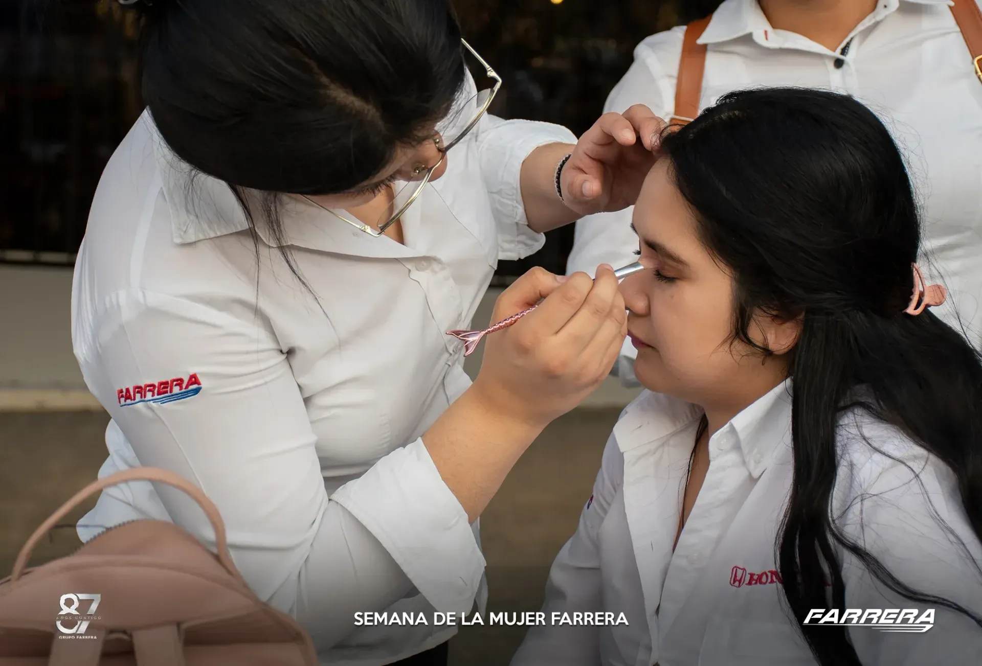 Maquilladora aplica maquillaje de ojos a una mujer sentada vestida de blanco durante una sesión de belleza.