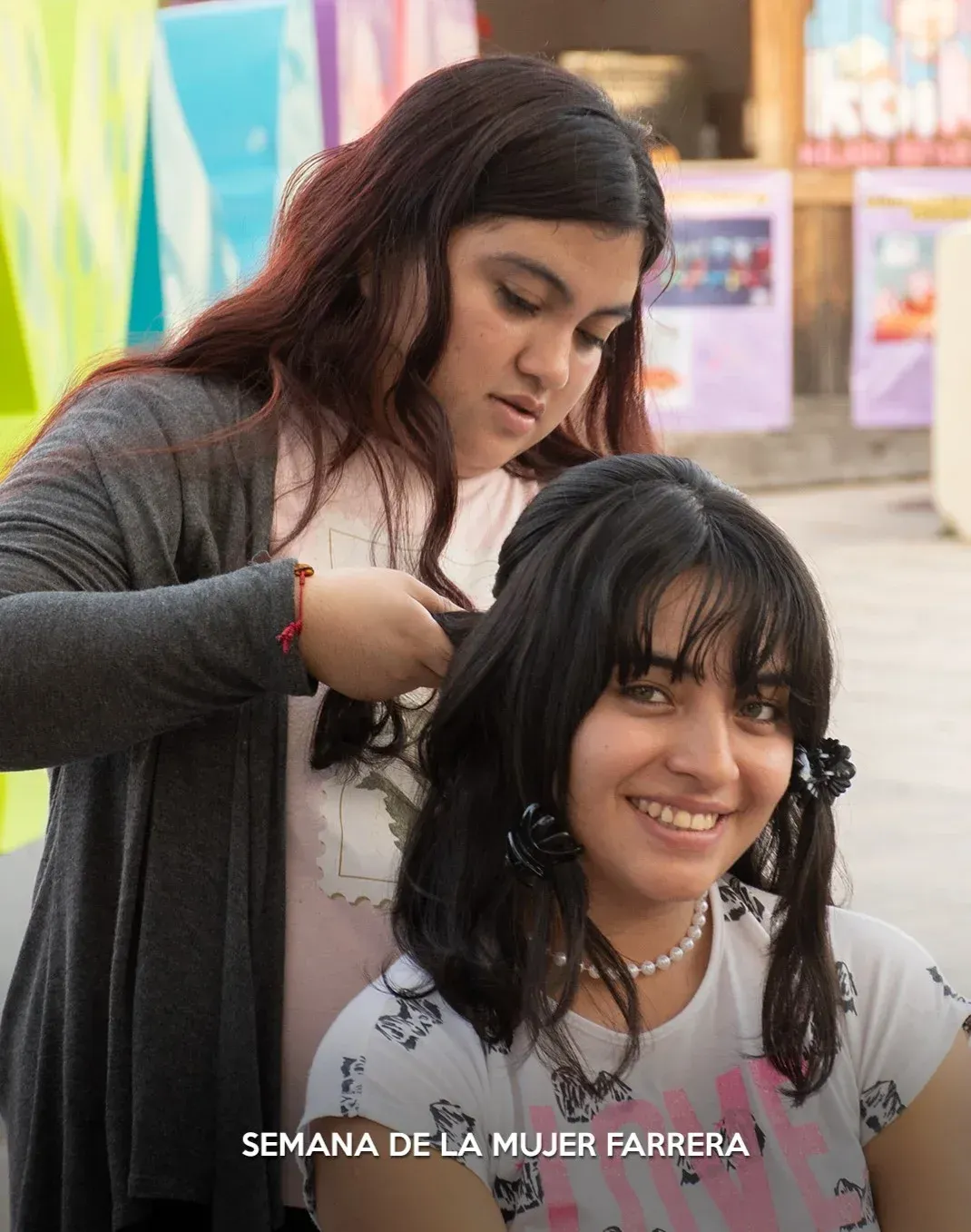 Mujer trenzando el cabello de otra mujer al aire libre; ambas sonriendo, con el texto “Semana de la Mujer Farera”.