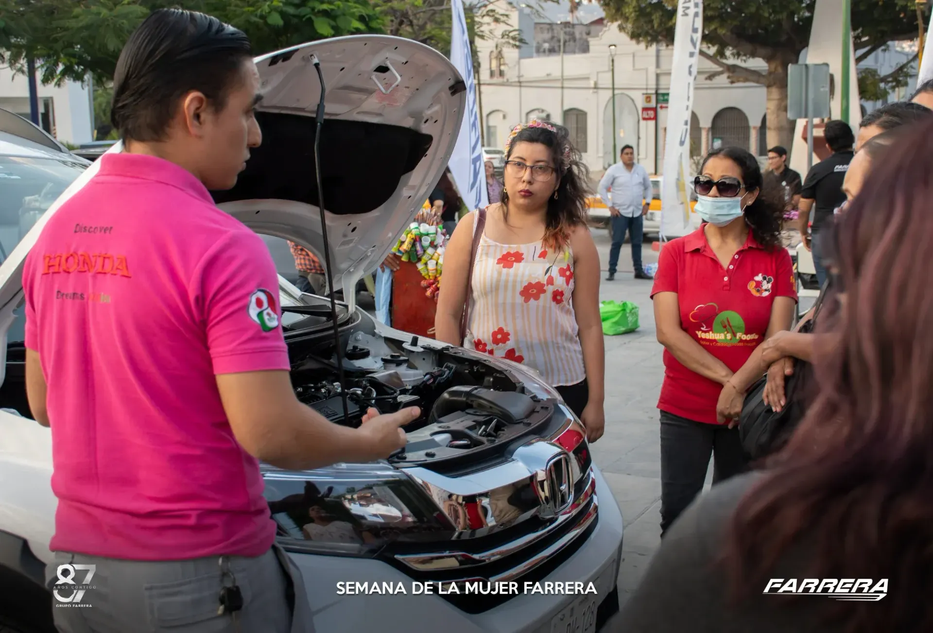 Varias personas inspeccionan un coche con el capó abierto en una calle de la ciudad; una de ellas muestra el motor.