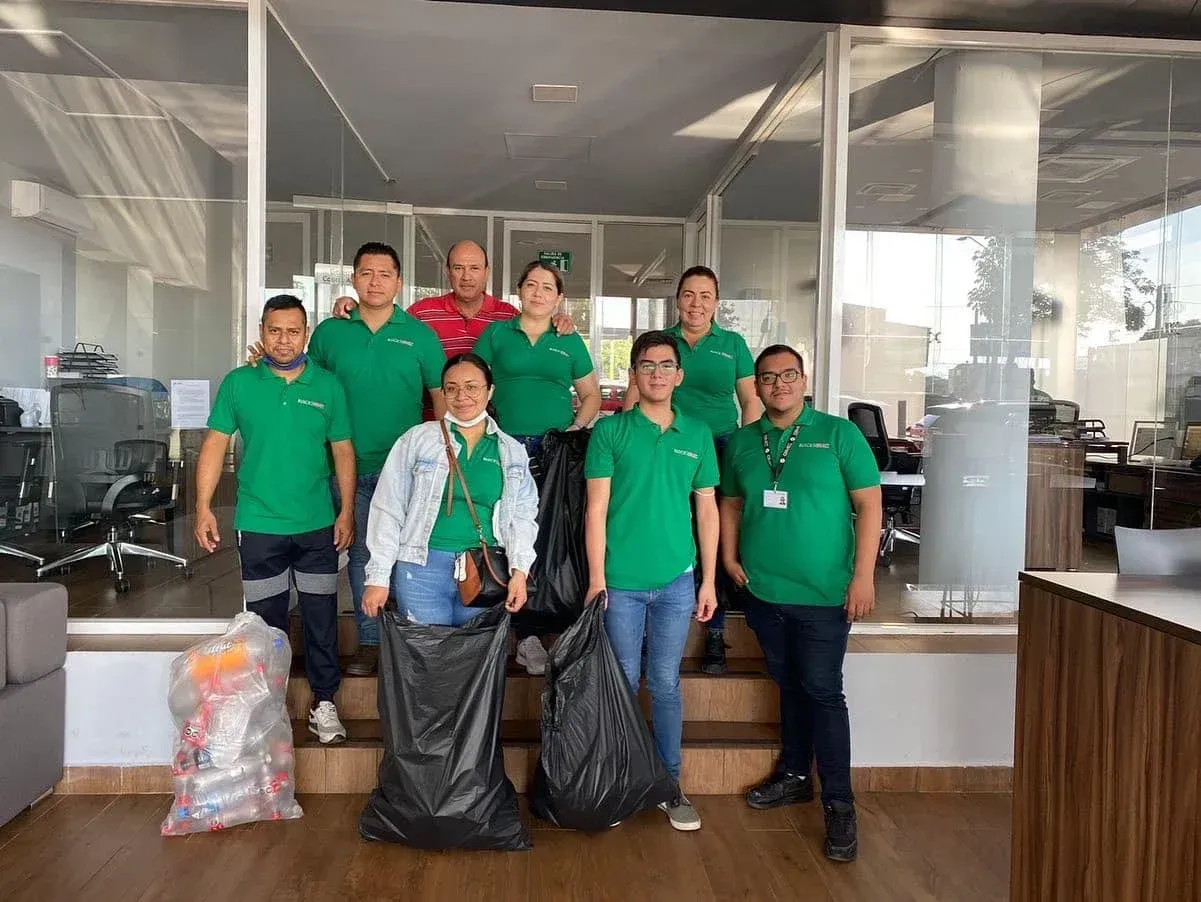 Equipo con camisetas verdes posando en el interior con bolsas de suministros frente a oficinas acristaladas.