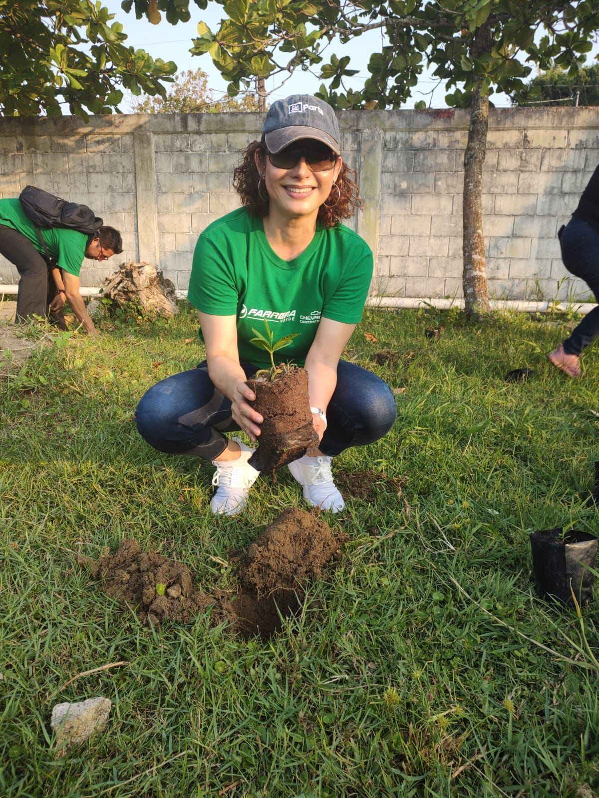 Mujer con camisa y gorra verdes plantando un árbol en un jardín, sosteniendo un cepellón y sonriendo.