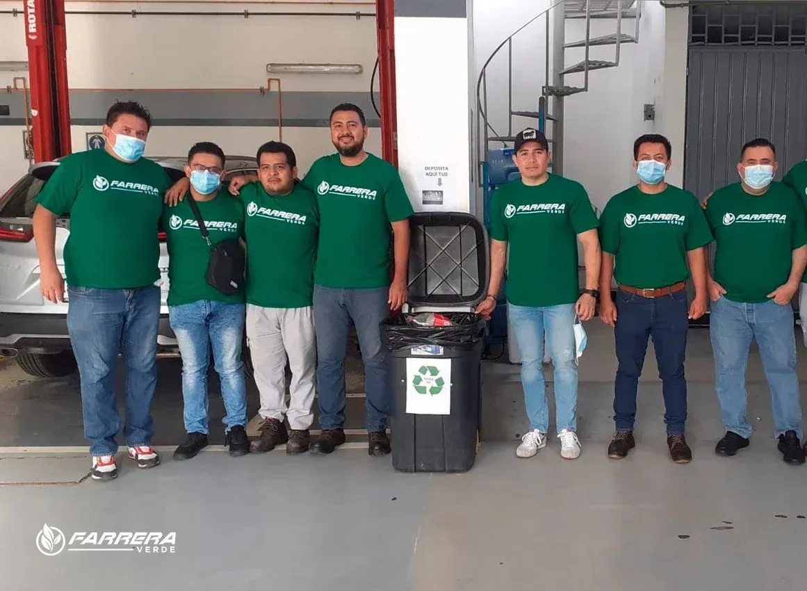 Seis personas con camisetas verdes están de pie en un garaje junto a un contenedor de reciclaje y las puertas de un camión.