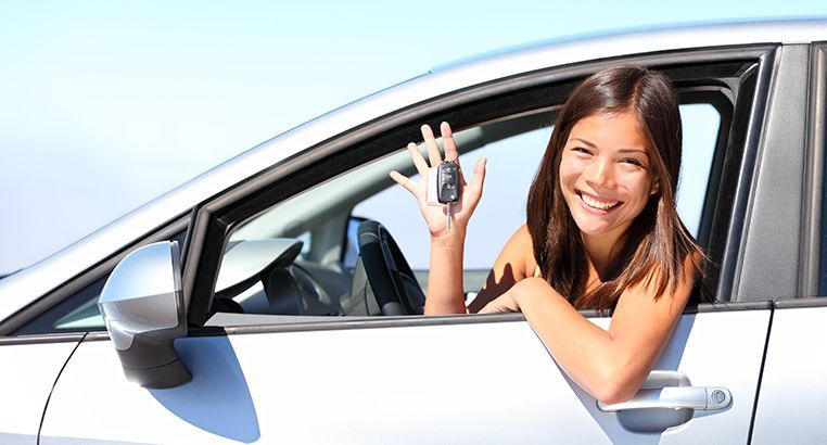 Mujer sonriente en un coche blanco sosteniendo las llaves del coche y saludando desde la ventanilla abierta.