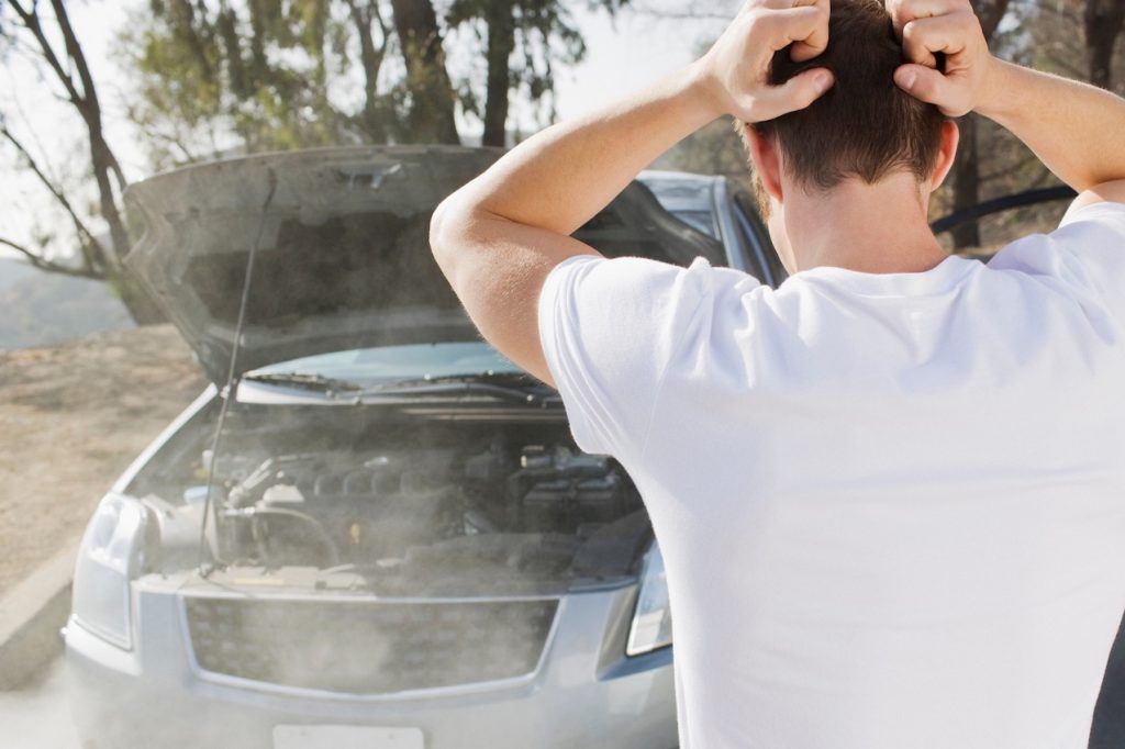 Una persona con camisa blanca mira dentro del capó abierto de un coche, del que sale vapor.