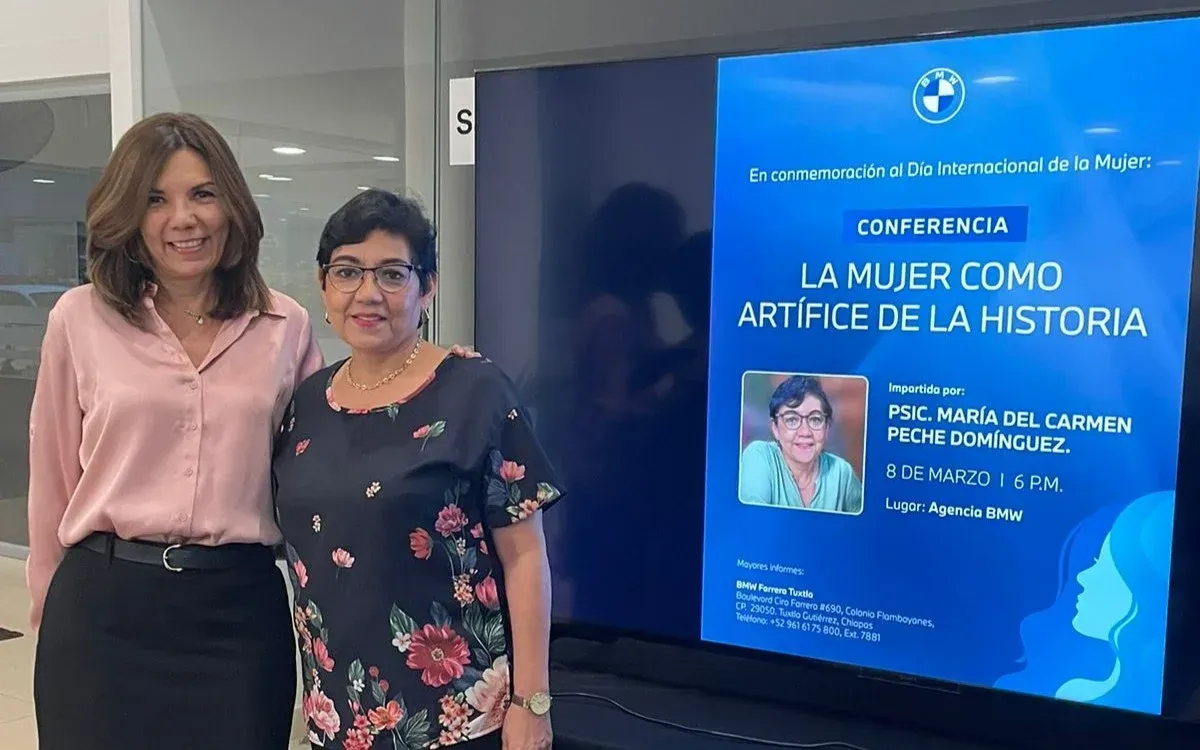 Dos mujeres de pie junto a un cartel azul de una conferencia en un pasillo interior.