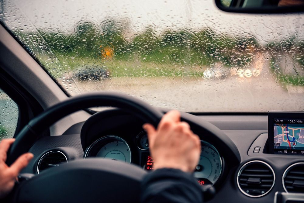 Vista del conductor a través de un parabrisas salpicado de lluvia, manos en el volante, tablero y GPS visibles.