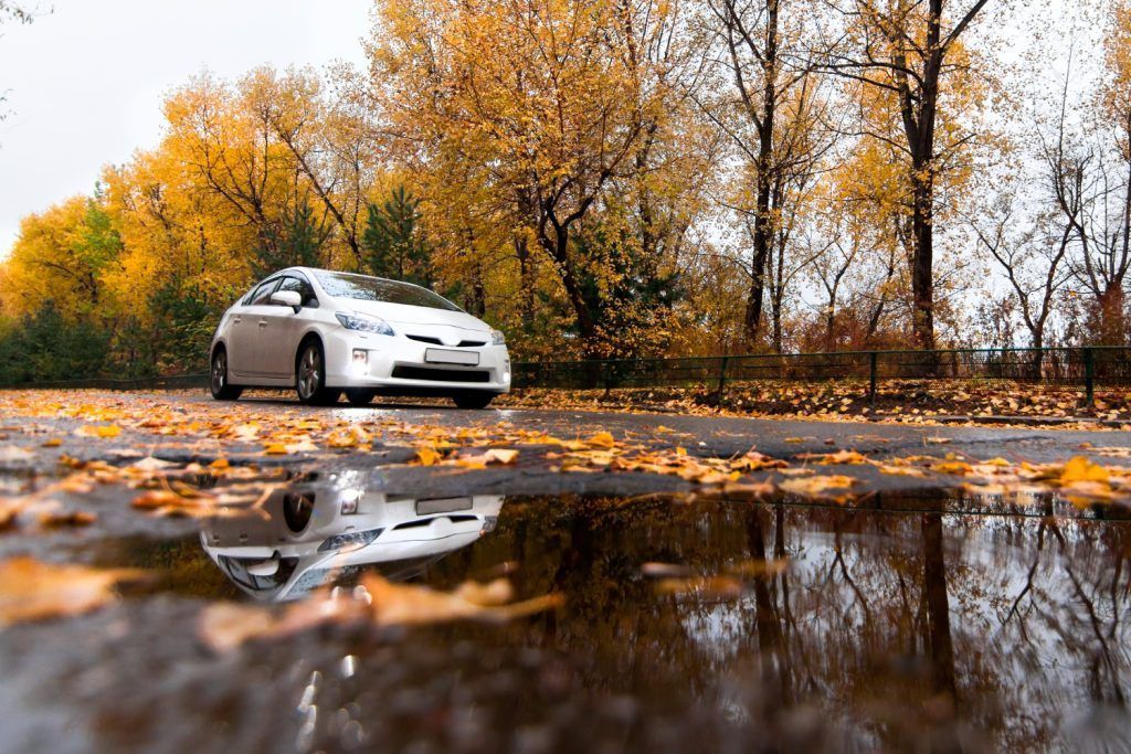Un coche blanco en una carretera mojada con árboles otoñales y su reflejo en un charco.