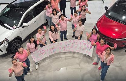 Un grupo de personas sostiene una pancarta grande entre un coche blanco y un coche rojo en una sala de exposición