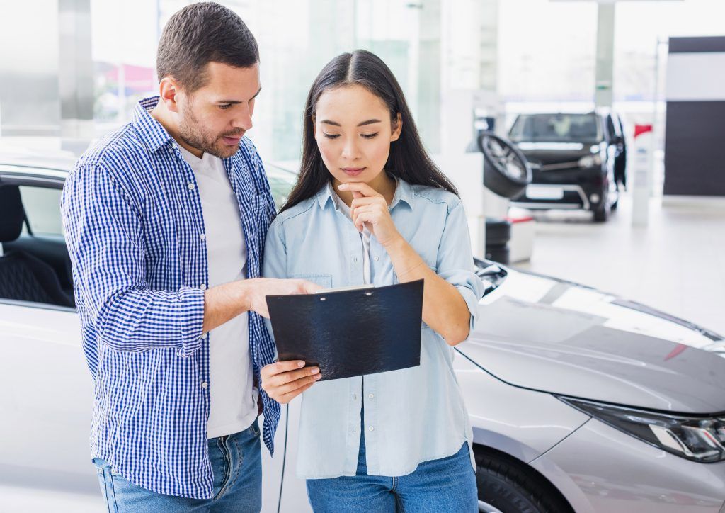 Dos personas revisan un documento junto a un coche plateado en una luminosa sala de exposición.