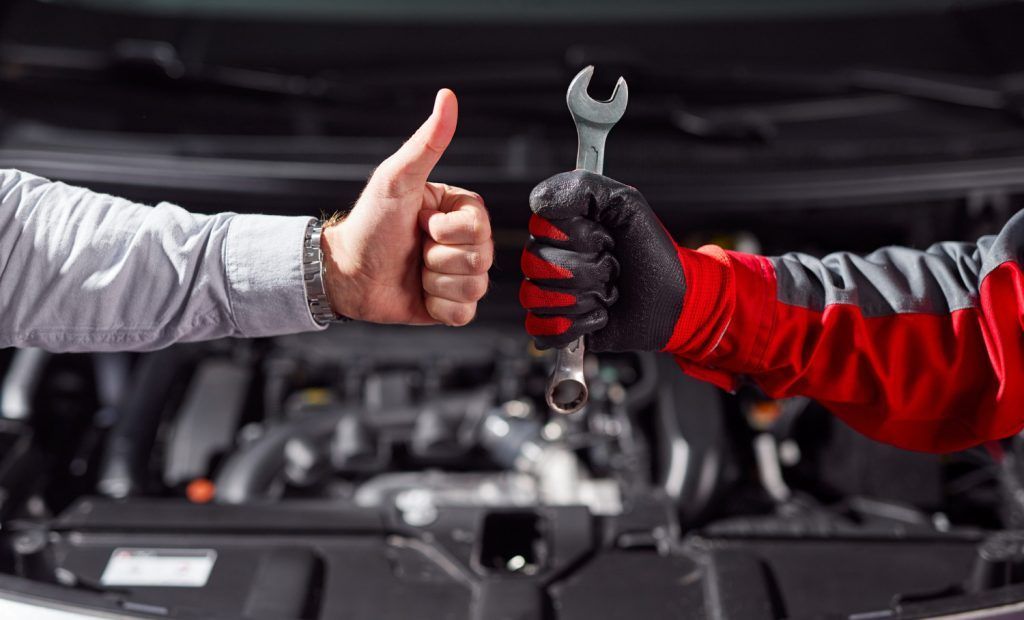 Dos mecánicos con guantes, uno levantando el pulgar y el otro sosteniendo una llave inglesa delante de un motor.