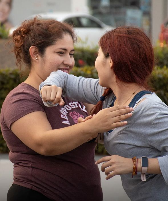 Dos mujeres sonriendo y abrazándose al aire libre, una con una camisa marrón y la otra con una blusa gris.